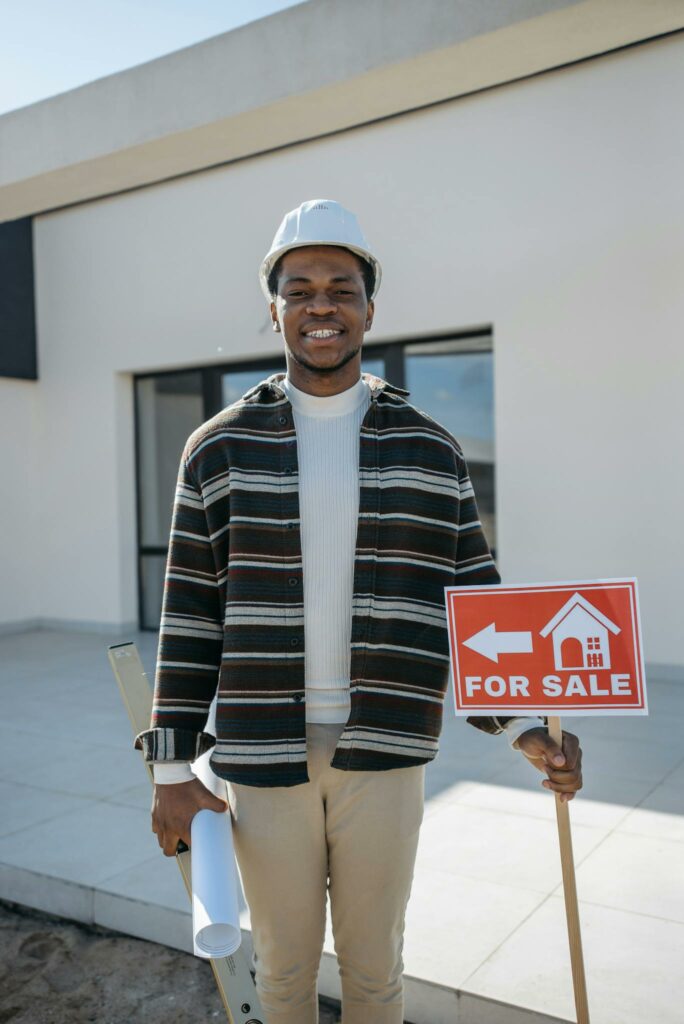 African American architect with a hard hat holding a 'For Sale' sign outside a building.